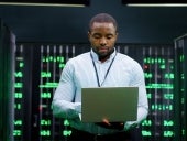 Male system administrator of big data center typing on laptop computer while working in server room. Programming digital operation. Man engineer working online in database center. Telecommunication.