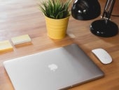 Closed MacBook on a desk with two sticky note pads, an Apple mouse, a desk lamp, and a small potted plan arranged around it.